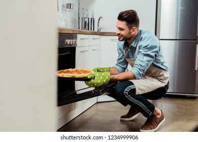 Smiling Young Man In Apron Taking Out Baking Tray With Pizza From Oven