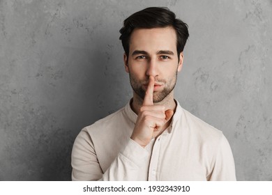 Smiling young man asking for silence gesturing with his finger isolated over gray background