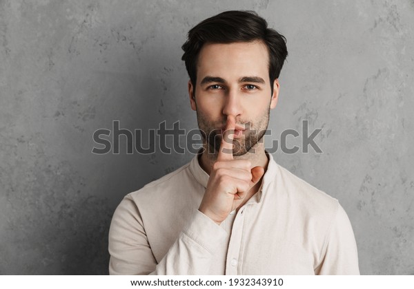 Smiling young man asking for silence\
gesturing with his finger isolated over gray\
background