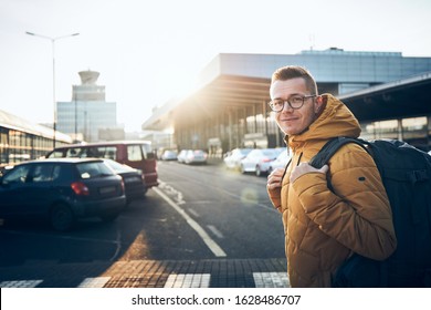 Smiling Young Man With Backpack On Car Park Aganist Airport Terminal Building At Sunset.