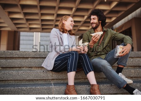Smiling young man and beautiful woman eating sandwich while in a conversation sitting on stairs