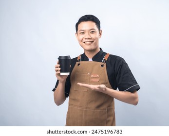 Smiling young man in a black uniform and brown apron, holding a black takeaway coffee cup and presenting it with his other hand against a plain light gray background - Powered by Shutterstock
