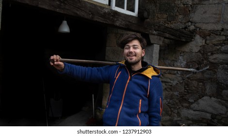 Smiling young man with blue clothes holding a rake in front of a stone house in a rural setting - Powered by Shutterstock