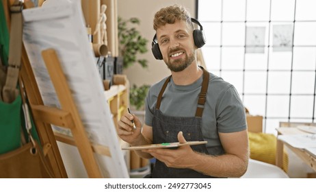 A smiling young man with blue eyes and a beard, wearing headphones, paints on a canvas in an art studio. - Powered by Shutterstock