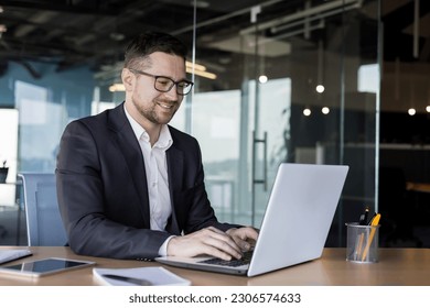 A smiling young man businessman in a business suit sits and works in the office on a laptop online, texts, chats with partners, clients. - Powered by Shutterstock