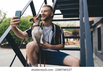 Smiling young man in casual clothes sitting on stairs with adorable Jack Russell Terrier in hands and taking self portrait on smartphone in city - Powered by Shutterstock