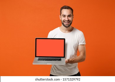 Smiling Young Man In Casual White T-shirt Posing Isolated On Bright Orange Background Studio Portrait. People Lifestyle Concept. Mock Up Copy Space. Holding Laptop Pc Computer With Blank Empty Screen