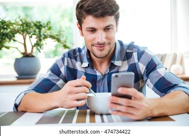 Smiling young man in checkered shirt using cell phone and having breakfast on the kitchen - Powered by Shutterstock