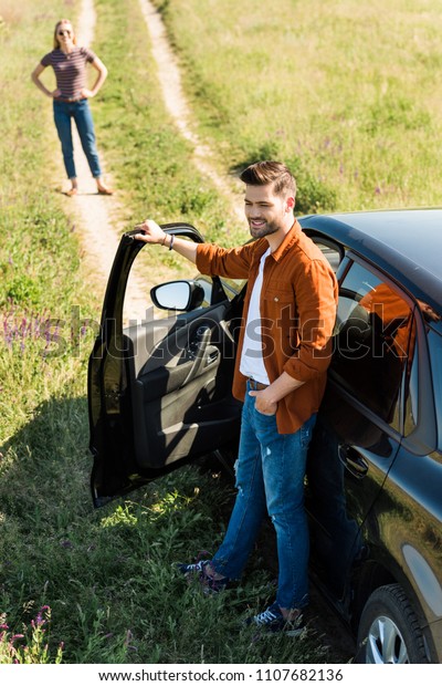 smiling young man closing door of car and\
his girlfriend waiting behind on rural meadow\
