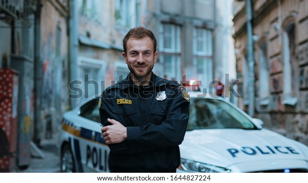 Smiling young man cops stand near patrol car look\
at camera wear glasses enforcement happy officer police uniform\
auto safety security communication control policeman close up slow\
motion