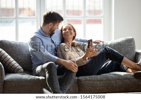 Smiling young man embracing lying on couch happy wife. Laughing millennial mixed race woman holding smartphone, showing funny video or photos to husband. Positive family couple relaxing on sofa.