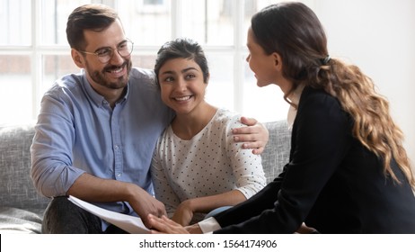 Smiling Young Man In Eyeglasses Hugging Pleasant Indian Wife, Listening To Confident Financial Expert Explaining Propose Details. Happy Family Couple Clients Meeting Professional Real Estate Agent.