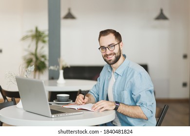 Smiling Young Man In Eyeglasses Sitting Alone At Table In Coffee Shop Cafe Restaurant Indoors Working Or Studying On Laptop Pc Computer Writing In Notebook. Freelance Mobile Office Business Concept