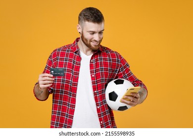 Smiling Young Man Football Fan In Red Shirt Cheer Up Support Favorite Team With Soccer Ball Using Mobile Cell Phone Hold Credit Bank Card Isolated On Yellow Background. People Sport Leisure Concept