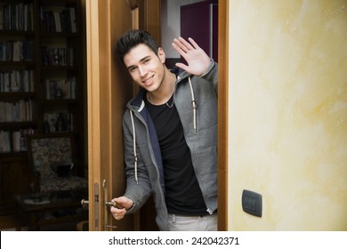 Smiling young man getting out of door waving at the camera with a friendly cheerful smile as he peers around the edge of a wooden door - Powered by Shutterstock