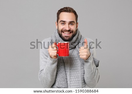 Smiling young man in gray sweater, scarf posing isolated on grey wall background. Healthy fashion lifestyle, cold season concept. Mock up copy space. Holding red cup of tea or coffee showing thumb up