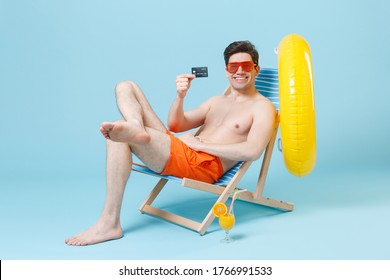 Smiling Young Man Guy In Orange Shorts Glasses Sit On Deck Chair Isolated On Pastel Blue Background Studio. People Summer Vacation Rest Lifestyle Concept. Mock Up Copy Space. Hold Credit Bank Card