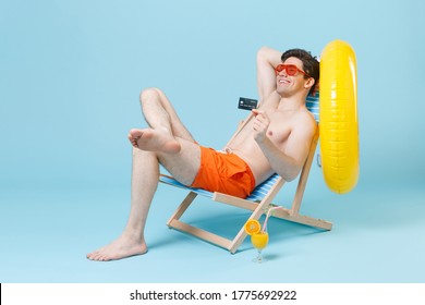 Smiling Young Man Guy In Orange Shorts Glasses Sit On Deck Chair Isolated On Pastel Blue Background Studio. People Summer Vacation Rest Lifestyle Concept. Mock Up Copy Space. Hold Credit Bank Card