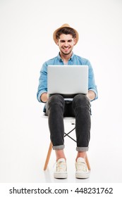 Smiling Young Man In Hat Sitting On Chair And Using Laptop Over White Background