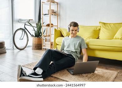 smiling young man with headphones on floor near yellow couch studying in laptop and writing in note