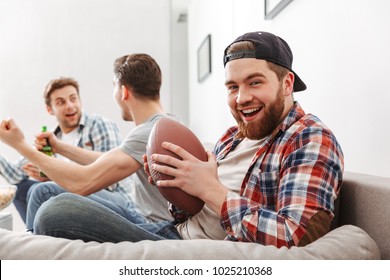 Smiling Young Man Holding American Football While Sitting With His Friends At Home On A Couch