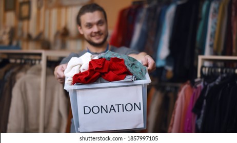 Smiling Young Man Holding Donation Box, Posing In Modern Charity Shop. Portrait Of Positive Guy Volunteer Working In Charity Organization And Sorting Clothes For Donation