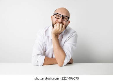 Smiling Young Man Holding Hand Under Chin While Sitting At Table On White Background.