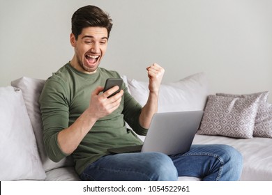 Smiling Young Man Holding Mobile Phone While Sitting On A Sofa At Home With Laptop Computer And Celebrating