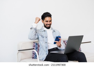 Smiling young man holding mobile phone while sitting on a sofa at home with laptop computer and celebrating