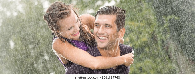 Smiling young man holding pretty girlfriend on his back during heavy summer rain