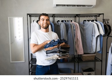 Smiling young man holding a stack of pants in his hands while standing in a walk in closet full of clothes