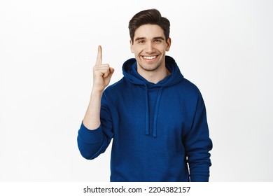 Smiling young man in hoodie, pointing finger up, showing advertisement, sale on top, standing over white background. - Shutterstock ID 2204382157