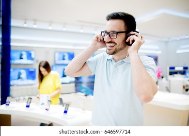 Smiling Young Man Listening To The Music In Tech Store. Trying On Headphones.