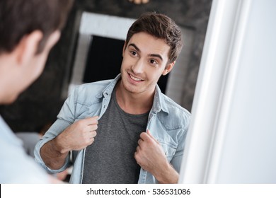 Smiling young man looking at himself in a mirror and holding his open shirt with two hands