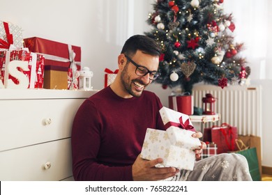 Smiling Young Man Opening Christmas Gift At Home