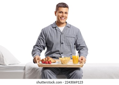 Smiling young man in pajamas sitting on a bed with a breakfast tray isolated on white background