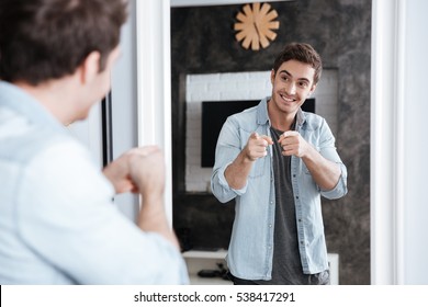 Smiling young man pointing fingers at his mirror reflection while standing at home