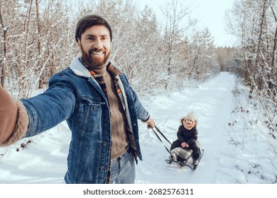 Smiling young man pulling sled with his daughter and making selfie