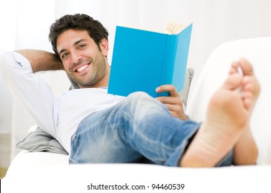 Smiling Young Man Reading A Book And Relaxing On Sofa At Home