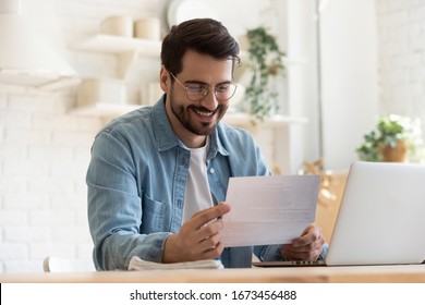 Smiling young man reading good news in paper notification, sitting at table with laptop, happy satisfied male wearing glasses holding document, job promotion, loan approval, great exam result