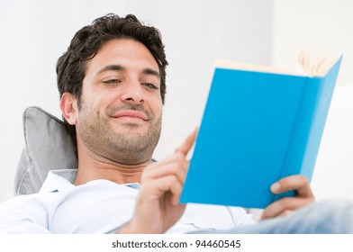 Smiling Young Man Reading A Novel Book While Relaxing On Sofa