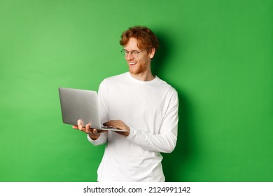 Smiling Young Man With Red Hair, Wearing Glasses, Working On Laptop And Smiling, Standing Over Green Background