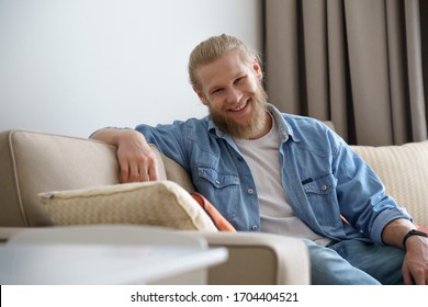 Smiling young man relaxing on sofa in modern apartment living room looking at camera, happy single millennial bearded hipster guy enjoying lifestyle lounge on comfortable couch at home alone. Portrait