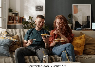 Smiling young man sharing french fries with his pretty girlfriend holding plate with hamburgers while both sitting on couch - Powered by Shutterstock