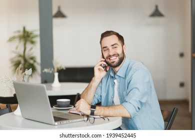 Smiling Young Man Sit At Table In Coffee Shop Cafe Working Studying On Laptop Computer Talking On Cell Phone Relaxing In Restaurant During Free Time Indoor. Freelance Mobile Office Business Concept