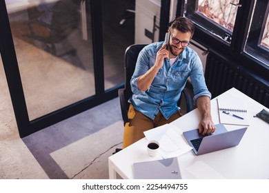 Smiling young man sitting in the office by the window and working on a laptop while talking on the phone. - Shutterstock ID 2254494755