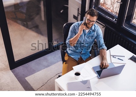 Smiling young man sitting in the office by the window and working on a laptop while talking on the phone.