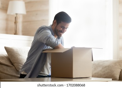 Smiling young man sitting on cozy couch at home, unpacking carton box with long-awaited purchased item. Happy male client satisfied with fast delivery service, unboxing order from internet store.