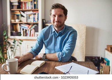 Smiling Young Man Sitting At A Table In His Living Room Writing Down Notes In A Notepad While Working From Home