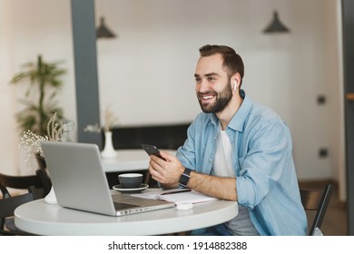 Smiling Young Man Sitting At Table In Coffee Shop Cafe Restaurant Indoors Working Studying On Laptop Pc Computer Listening Music With Air Pods Cell Phone. Freelance Mobile Office Business Concept